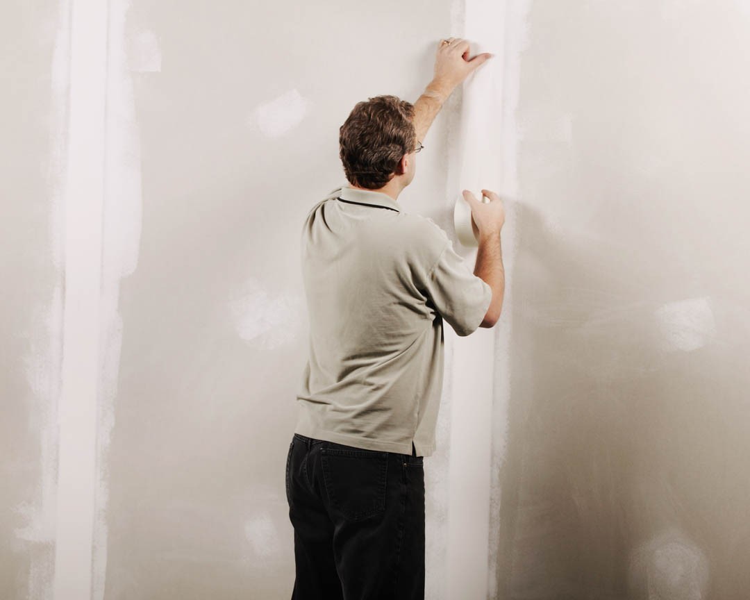 The image shows a man standing next to a wall with construction materials on it, working on a task that involves measuring or marking the surface.