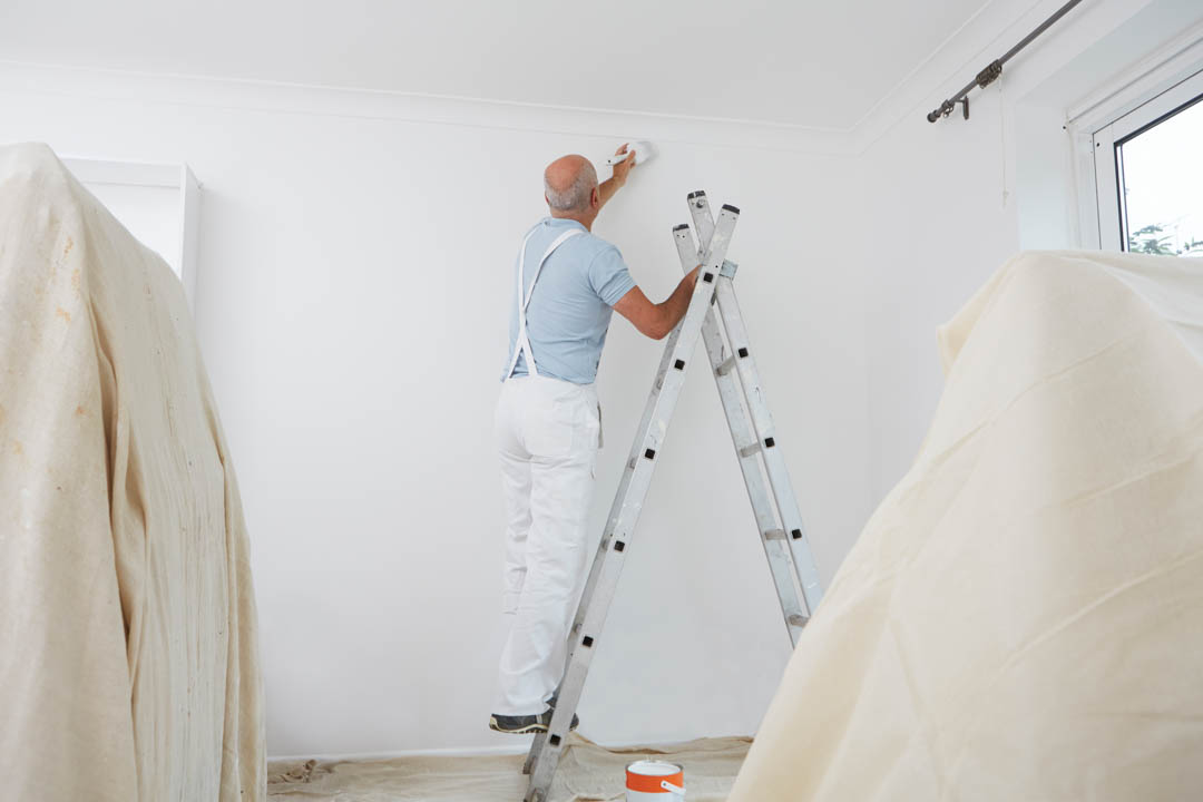 A person using a ladder to paint a wall in a room with white walls and plastic sheeting on the floor.