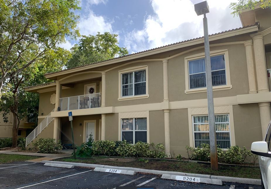 The image shows a two-story residential building with a beige exterior, featuring a balcony on the second floor, a garage door, and a driveway leading to a parking space.