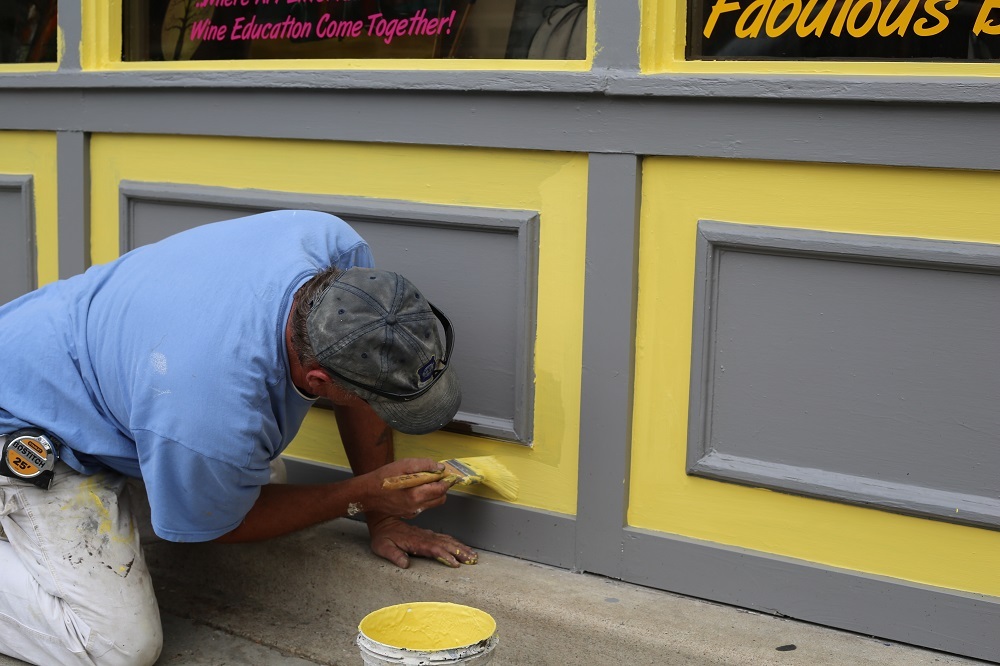 The image shows two individuals working on a yellow door frame with a paint bucket, likely engaged in painting or maintenance work, captured from an angle that emphasizes their activity.