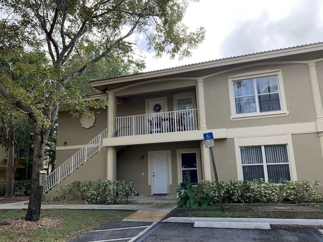 The image shows a two-story residential building with a beige exterior, white railings on the second floor balcony, and a tree partially visible in front of the structure.