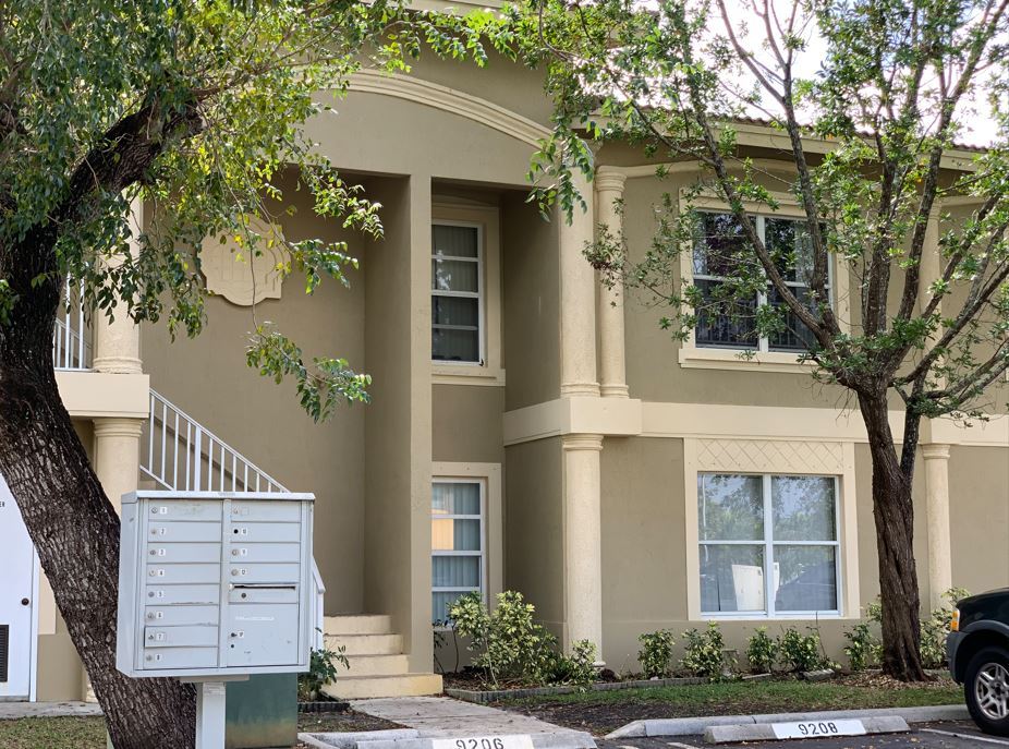The image shows a two-story residential building with beige walls, white trim around windows and doors, and a covered front porch with a staircase leading up to it. A mailbox is attached to the bottom of the front door. In front of the house, there's a tree with green leaves and a car parked on the street. The sky appears overcast.