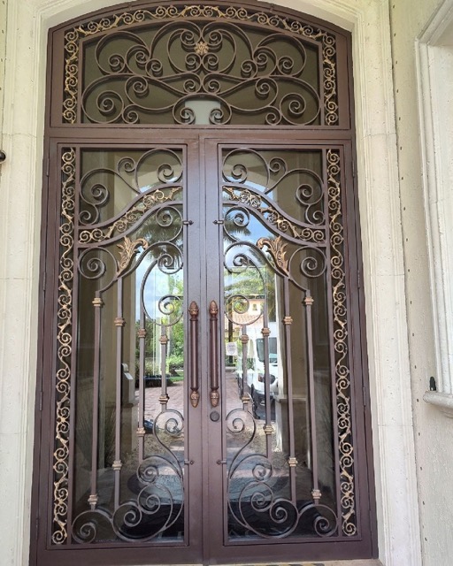 The image displays an ornate metal door with intricate scrollwork, set within a wooden frame, featuring decorative wrought iron elements and a large glass window at its center.