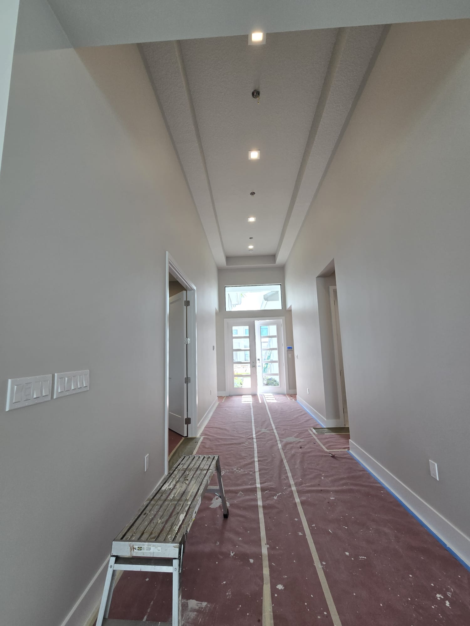 The image shows an interior hallway with a high ceiling, featuring recessed lighting and a long, narrow space leading towards a door at the end.