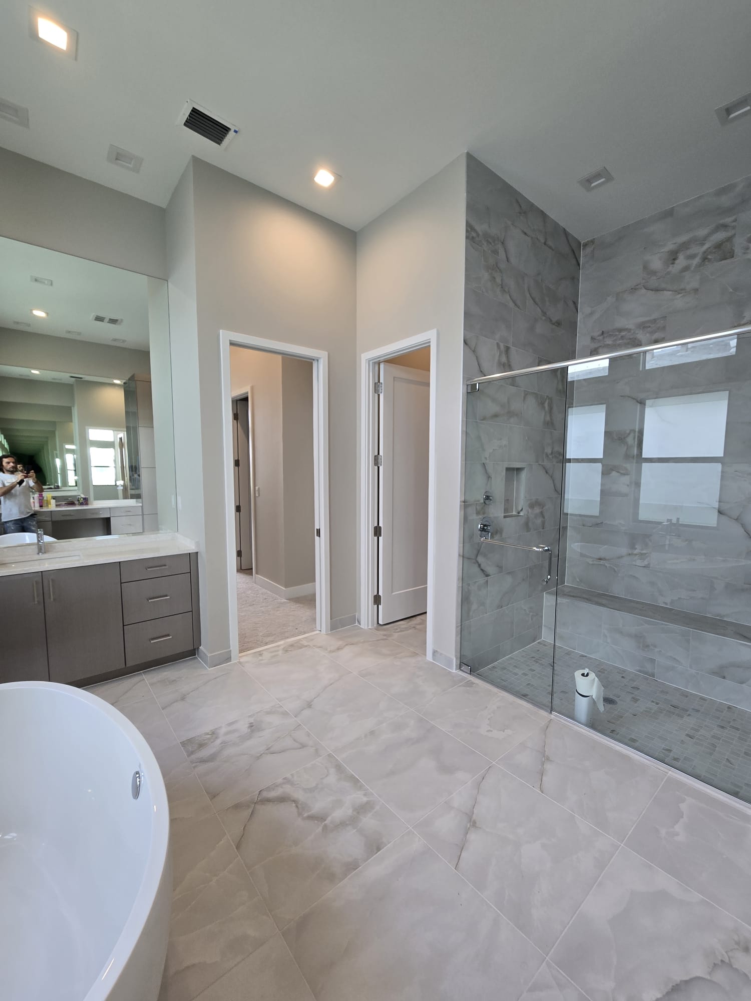 The image shows a modern bathroom with a beige tile floor, a large shower area with clear glass doors, a freestanding bathtub, and contemporary fixtures.