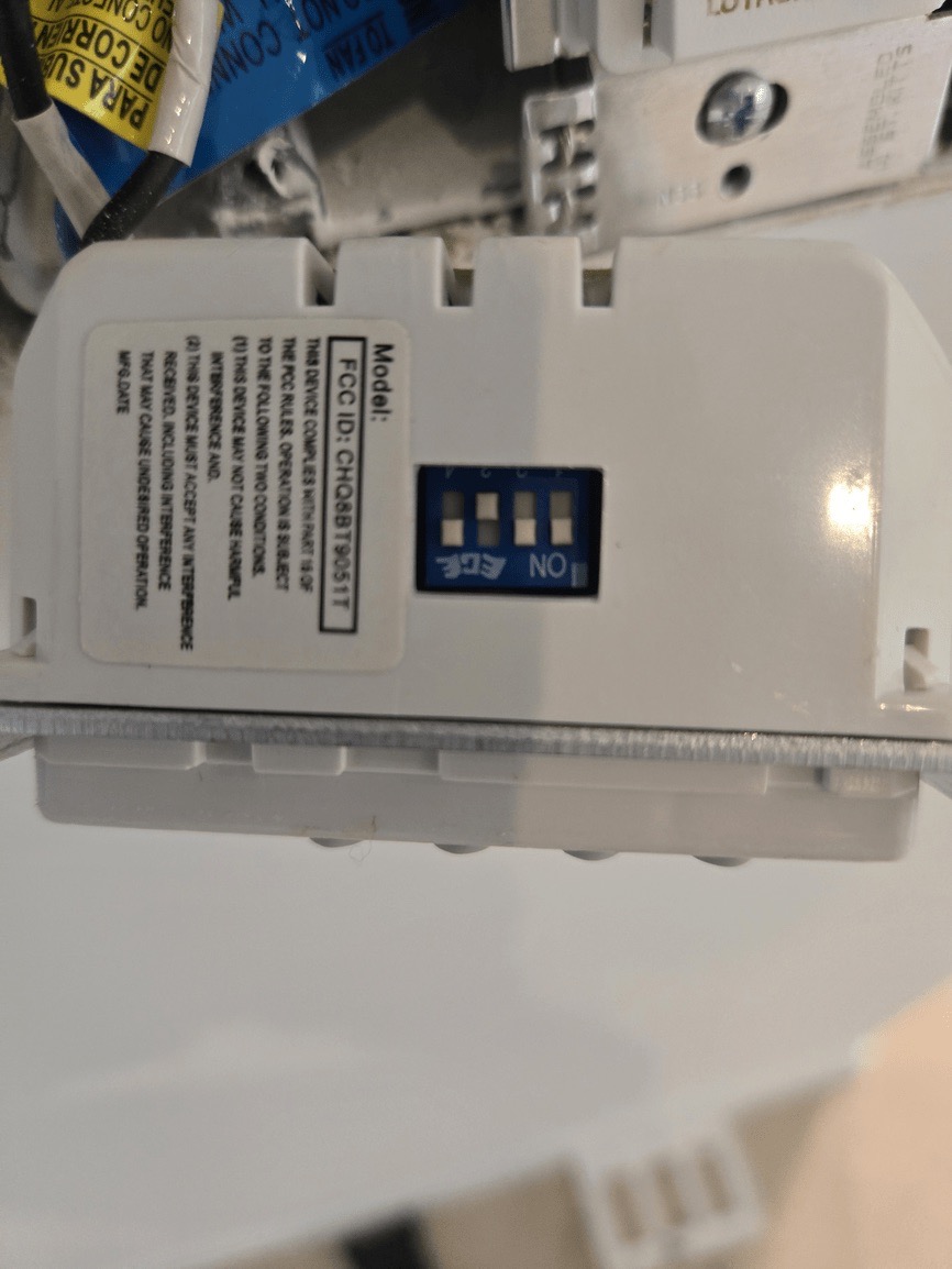 The image shows a white electronic device with a label on its side, featuring a barcode and text, against a blurred background that appears to be indoors.