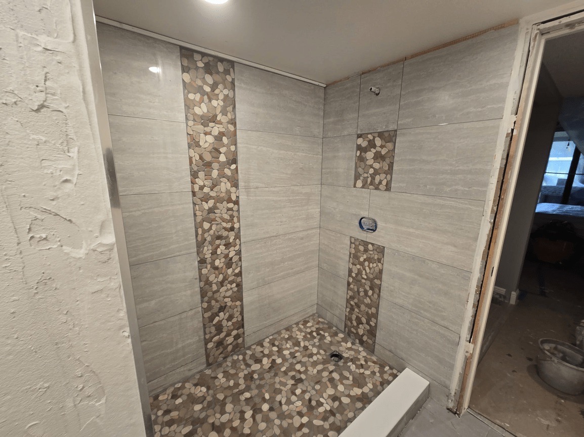 The image displays a bathroom interior featuring tiled walls with a stone-like pattern, a dark floor, and a shower area with multiple showerheads.