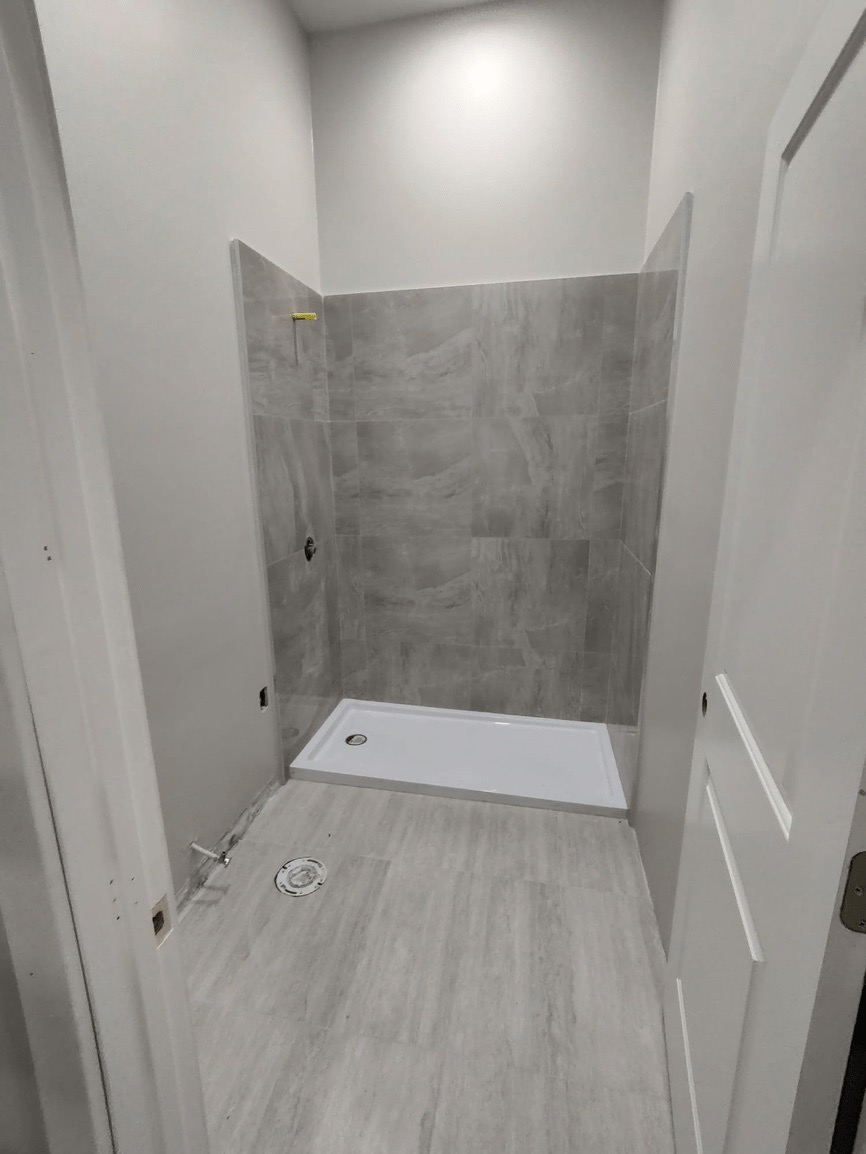 The image shows a small bathroom with a shower stall, a white floor, and a tiled wall.