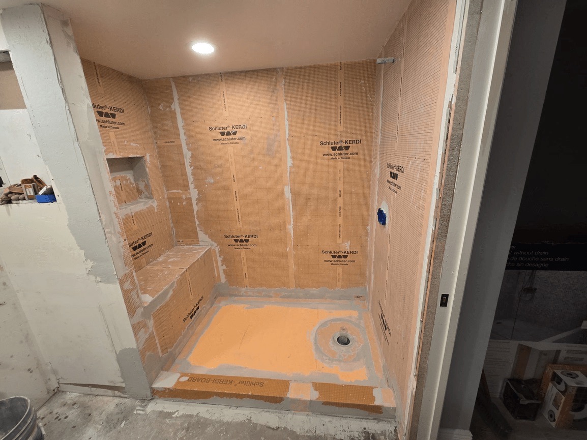 The image shows a bathroom undergoing renovation, with exposed walls and fixtures, including a shower area, and construction materials such as plywood and cardboard on the floor.