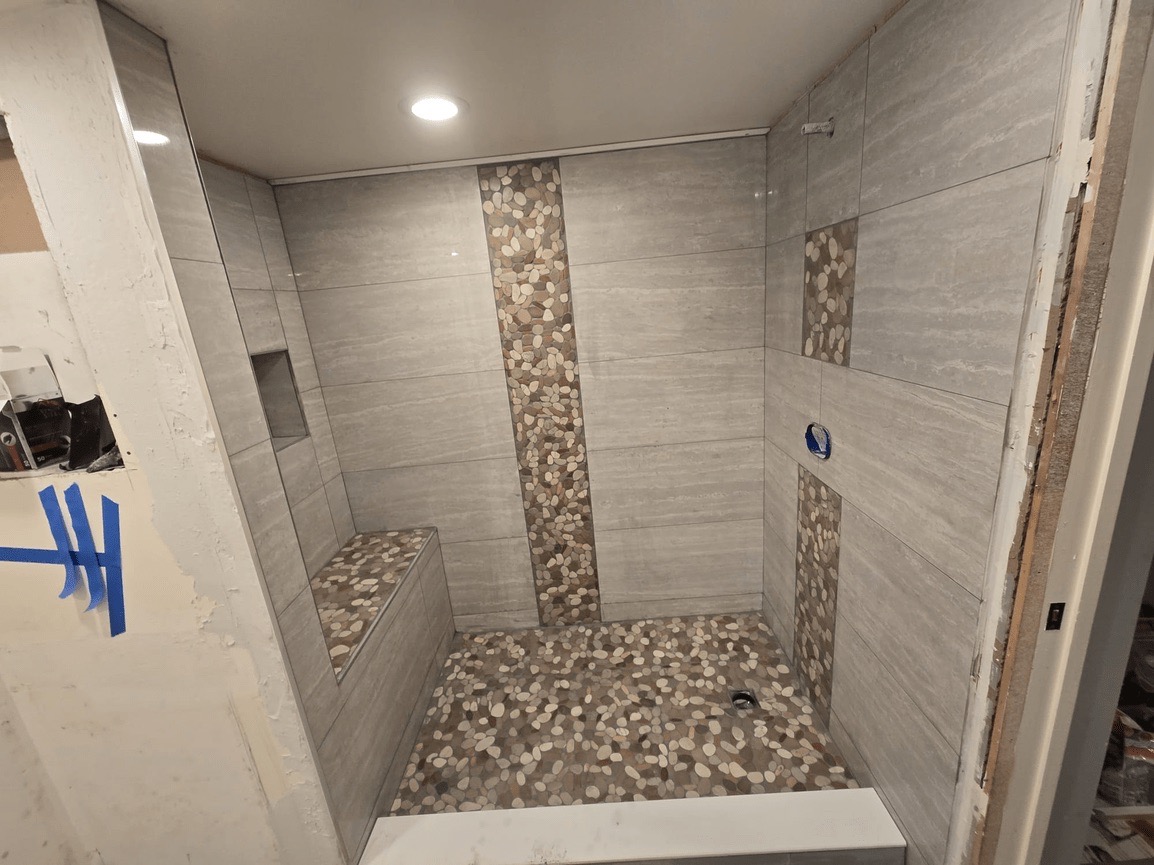 The image shows a bathroom under renovation with a partially tiled wall, a floor with small tiles, and a shower area with a bench seat.