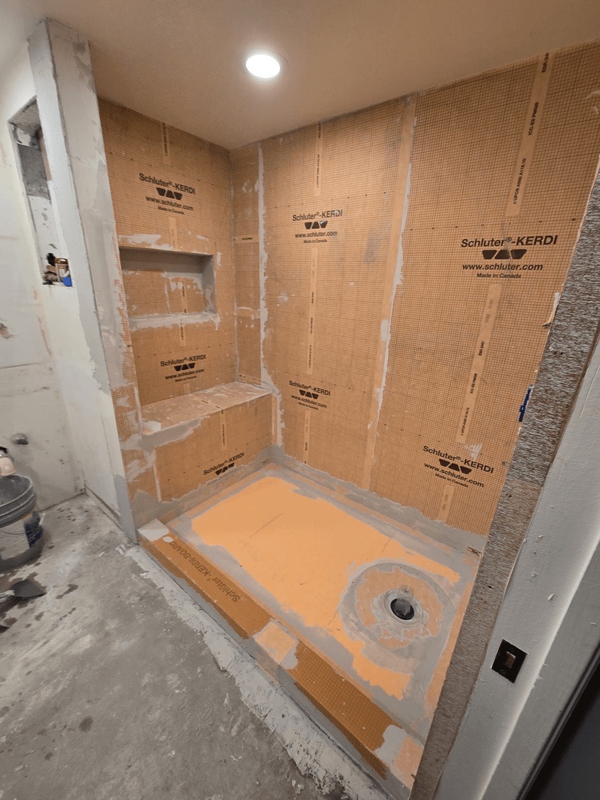 The image shows a bathroom under construction with exposed walls and floor, featuring cardboard sheeting and construction materials.