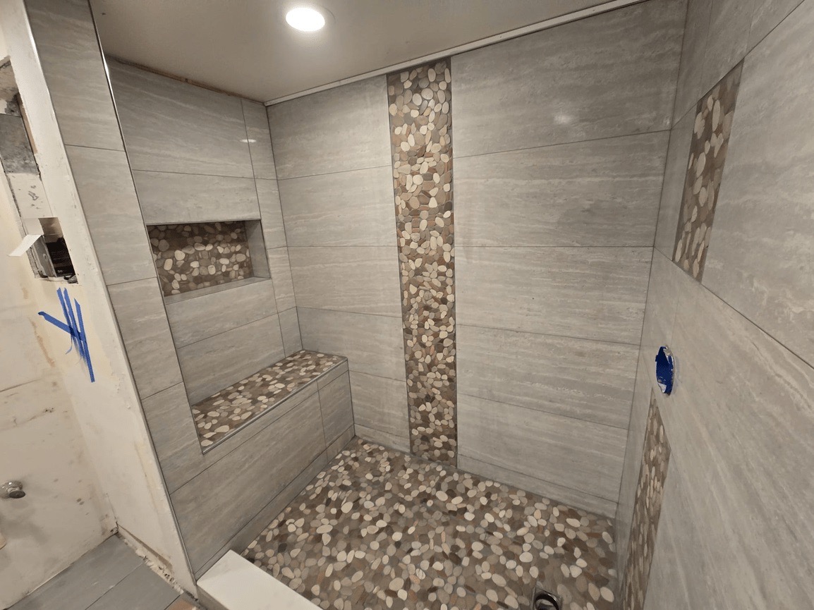 A small bathroom with a tiled floor and walls featuring a mix of marble-like tiles and stone-like accents, a shower stall with a glass door, and a bench inside.