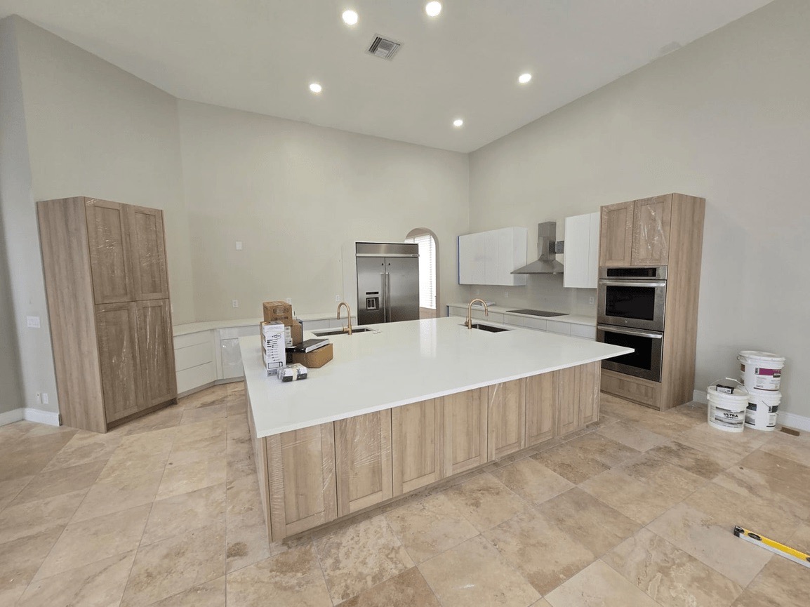 The image shows a modern kitchen interior with light-colored cabinets, a large island countertop, and a clean, white floor.