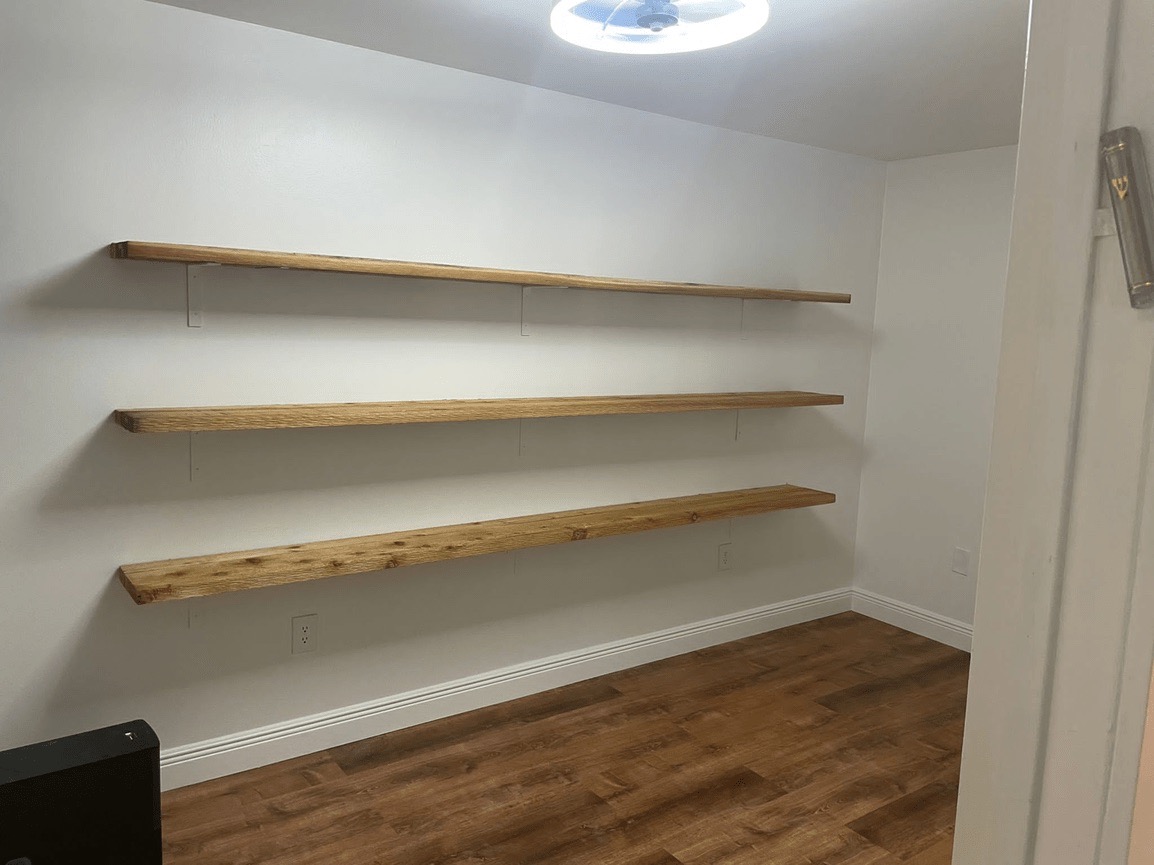 The image shows an interior space with wooden shelving units against a white wall, under a light fixture, with a dark-toned floor and a door frame visible on the right side of the photo.