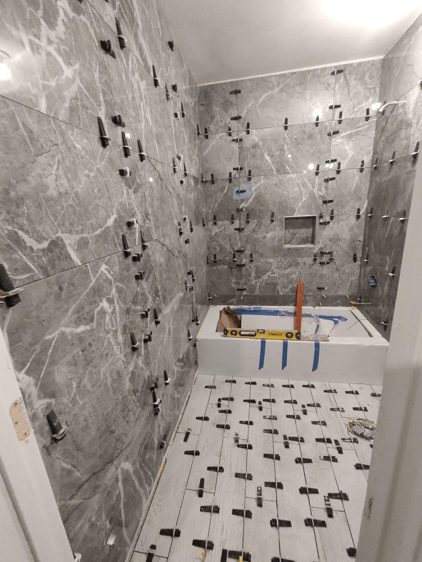 The image shows a bathroom under renovation with marble walls and floor, featuring exposed electrical wiring, pipes, and fixtures, including a sink and bathtub, and a partially installed wall-mounted cabinet.