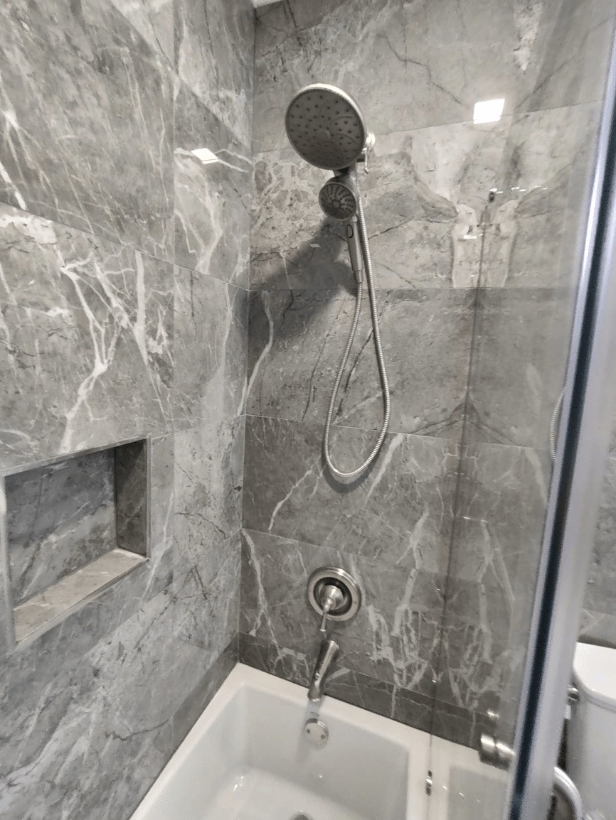 The image shows a modern bathroom with marble walls and flooring, featuring a walk-in shower with a glass door, a chrome faucet, and a towel rack inside the shower.