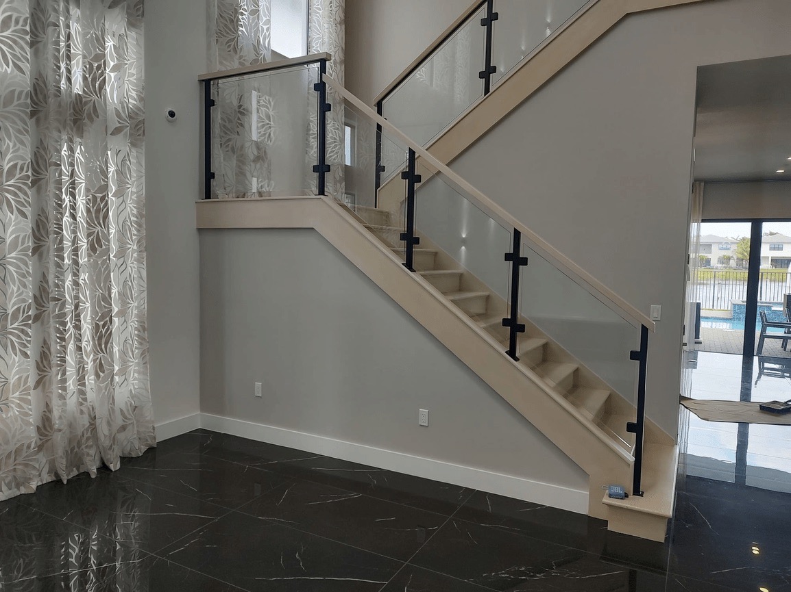 The image shows an interior view of a staircase with a wooden handrail leading up to a modern glass door, set against a backdrop of a room with a gray wall, a white curtain, and a dark floor.