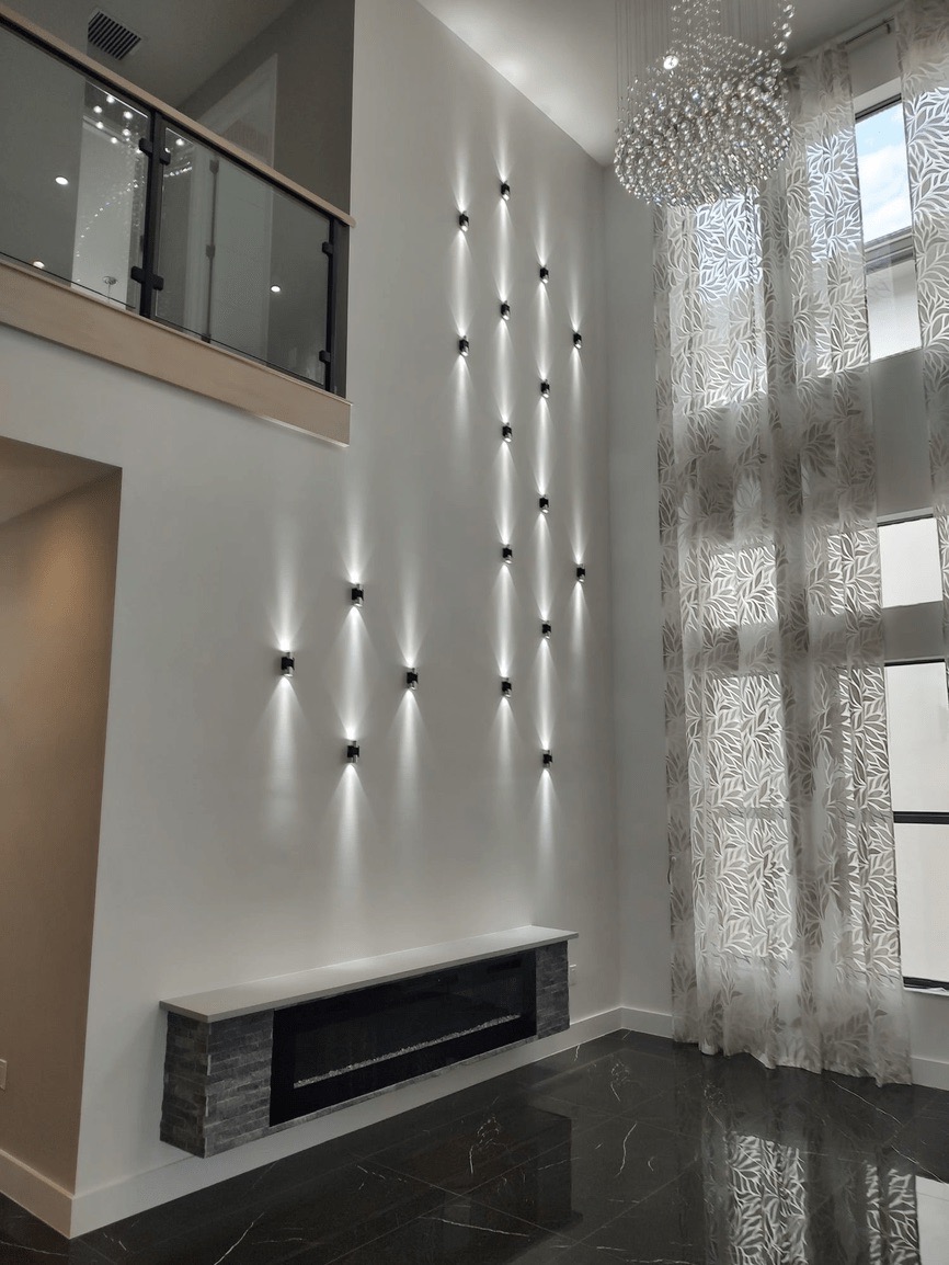The image shows a modern interior space with a contemporary fireplace, a striking wall decoration composed of vertical light fixtures, and a clean, minimalist aesthetic.