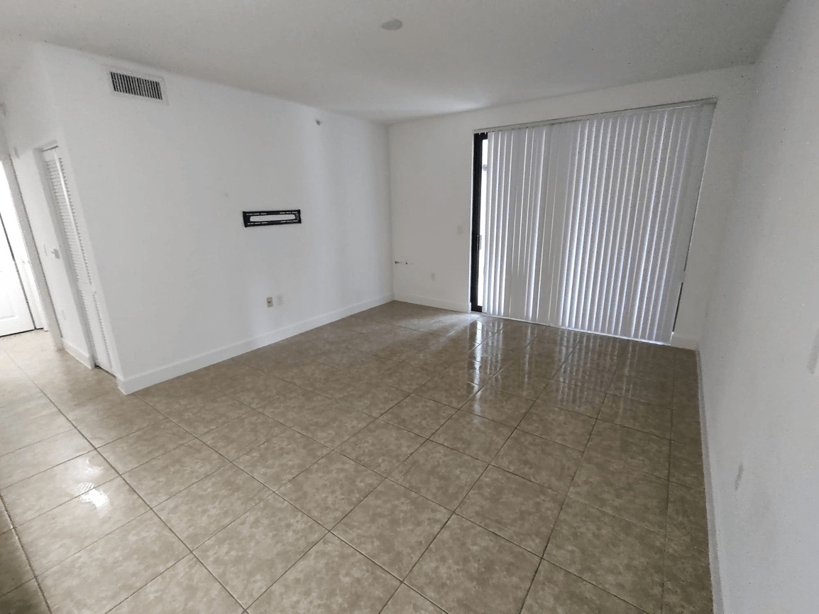 The image shows an interior space of a room with white walls, a tiled floor, and a large window covered by vertical blinds.