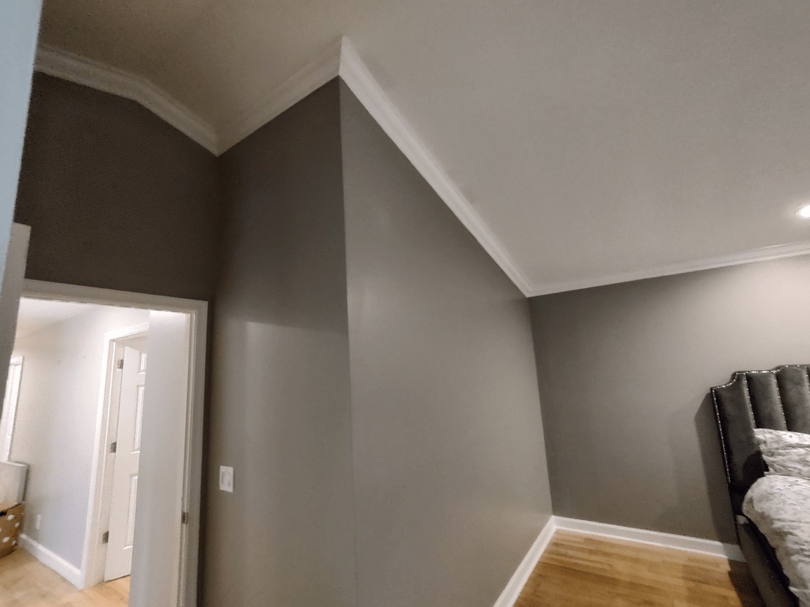 The image shows an interior space with a dark gray wall, a white ceiling, a wooden floor, and a corner where a wall meets another wall at a 90-degree angle. There's a glimpse of a bedroom area on the right side. The photo appears to be taken from a doorway or hallway entrance.