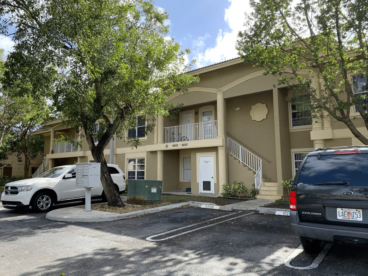 The image depicts a multi-story residential building with a beige exterior, situated within a parking lot with several vehicles parked nearby.
