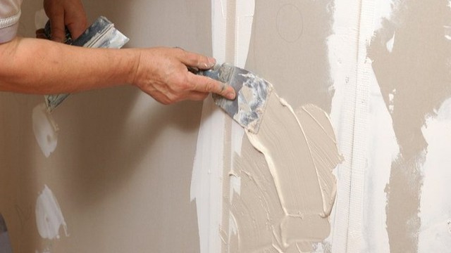 The image shows a person applying wallpaper to a wall using a brush and a spackling knife.