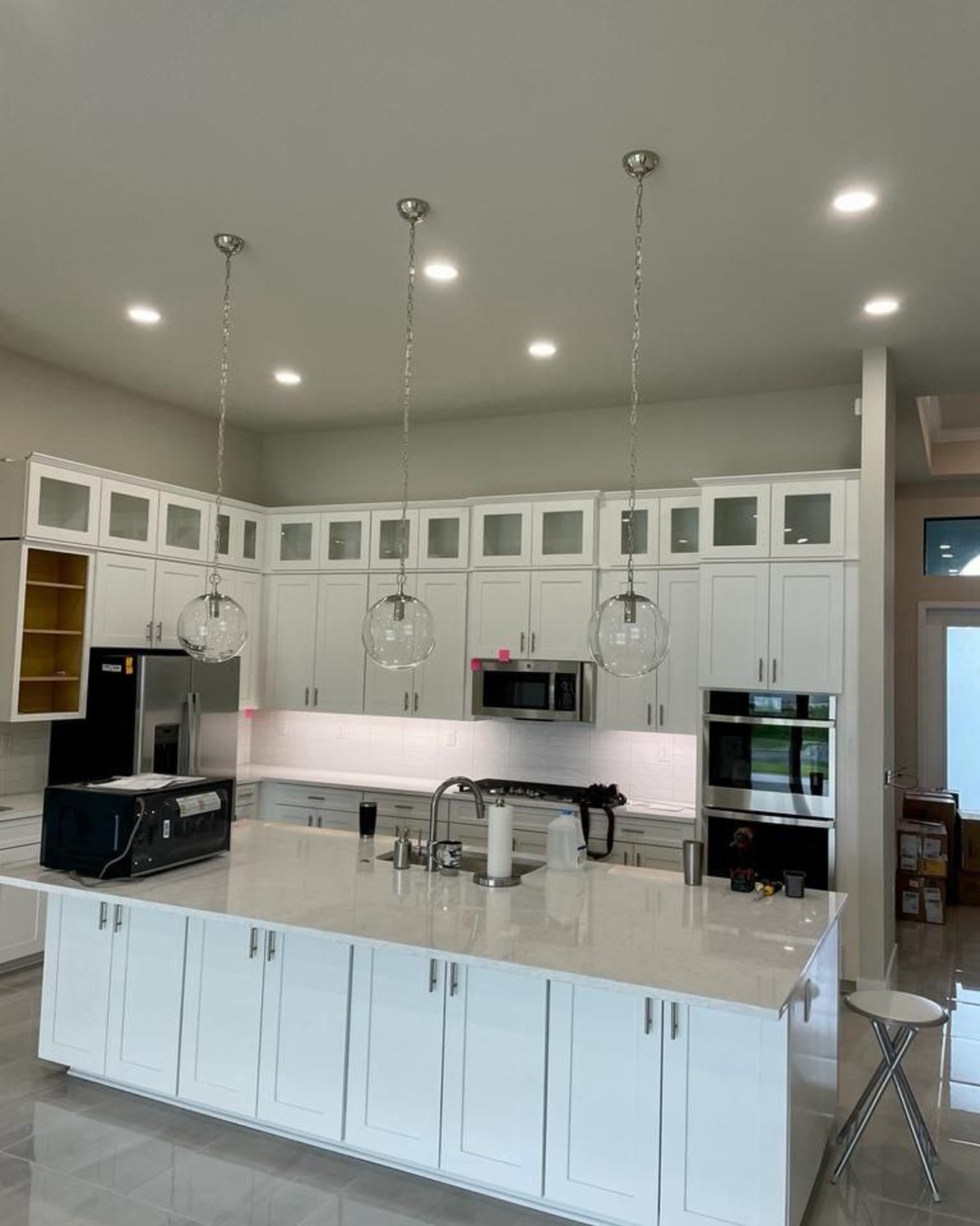 The image shows a modern kitchen interior with white cabinets, a large island countertop, stainless steel appliances including a refrigerator, stove, and microwave, a sink, and pendant lights above the island.
