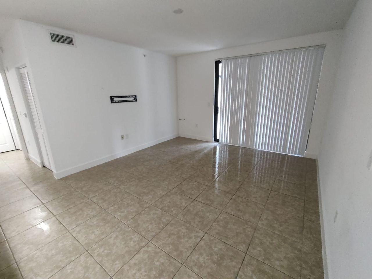 The image shows an interior view of a room with tiled flooring, closed white blinds on windows, and sliding glass doors leading to another space.