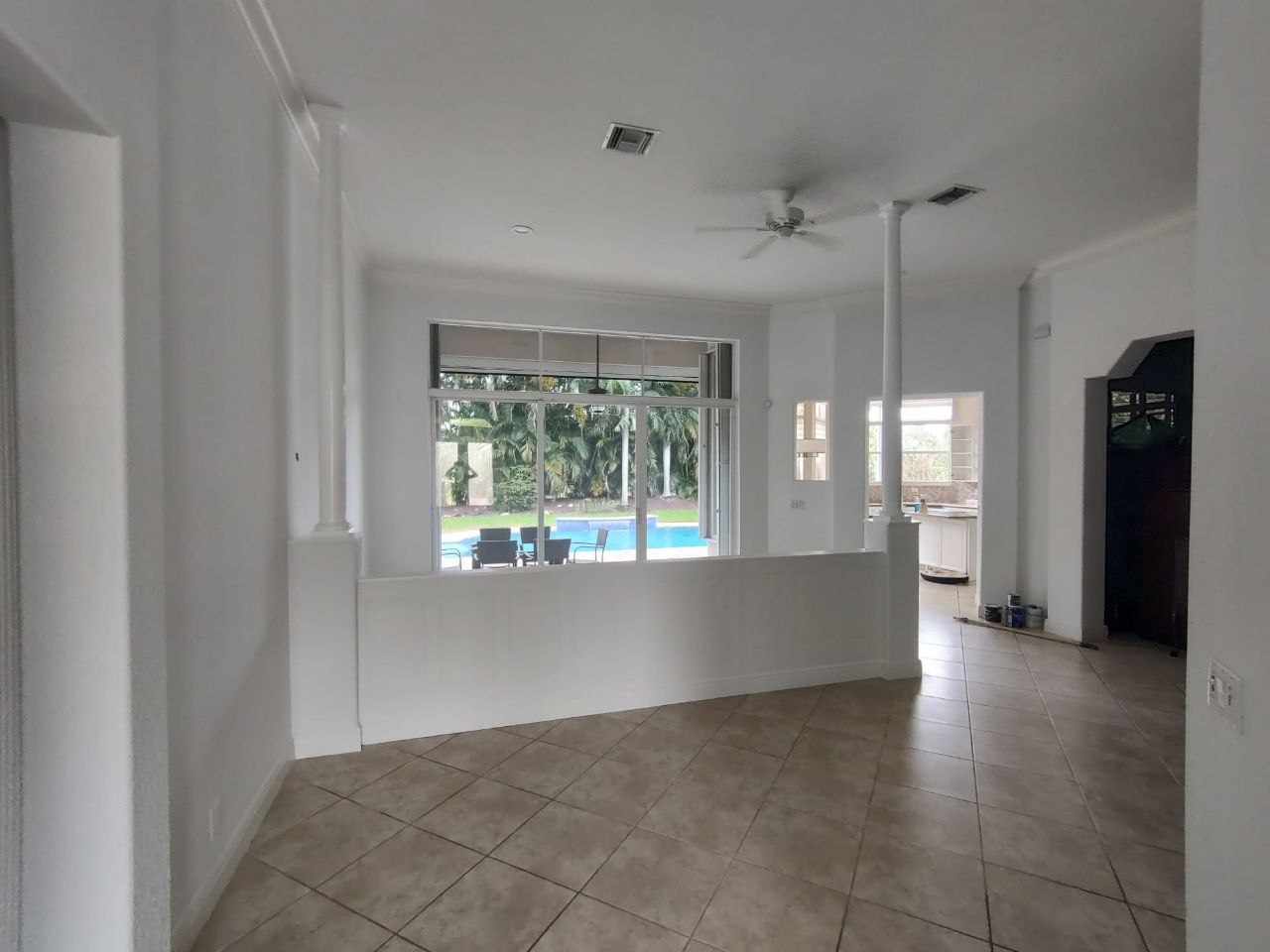 The image shows an interior view of a room with white walls and tiled flooring, featuring a large open space with a kitchenette to the left and a dining area with a table and chairs visible in the center.