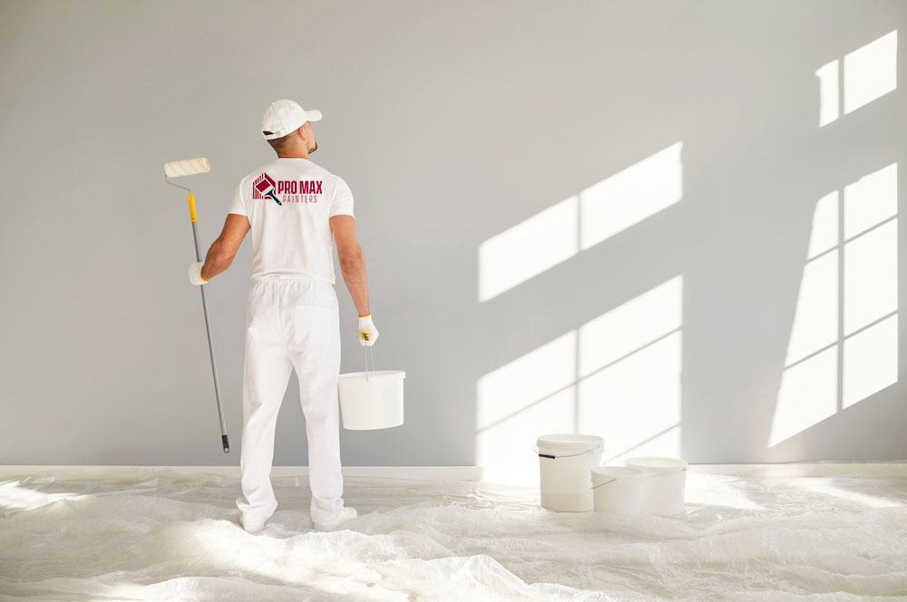 The image shows a person wearing white protective clothing, including gloves and goggles, holding a spray bottle, standing in front of a wall that appears to be under construction or renovation, with sunlight streaming through an open window.