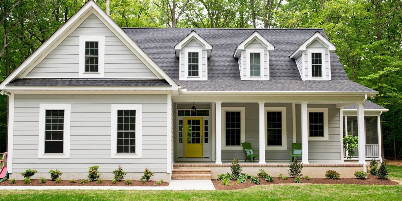 The image shows a two-story house with white siding, black shutters, a shingled roof, and a front porch. It has multiple windows and a garage with a white door.