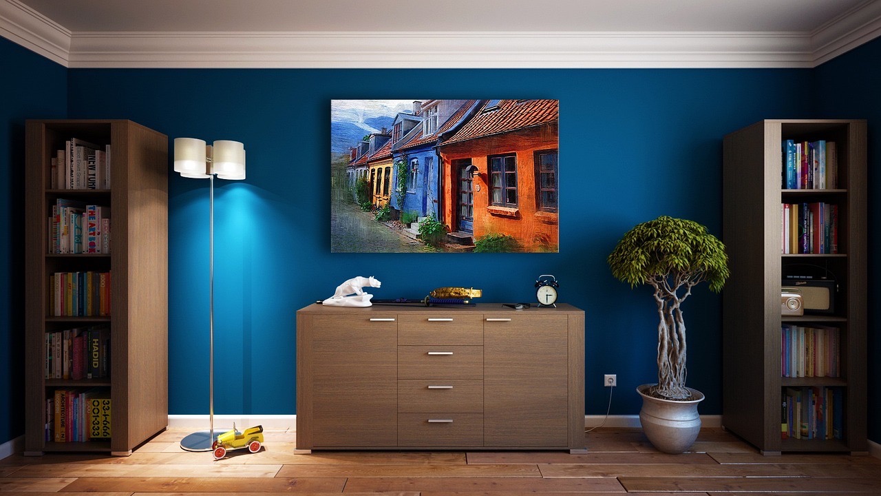 A living room scene with a modern blue wall, wooden floor, a large window, a bookshelf, a TV stand, a flat-screen TV, a beige sofa, a coffee table, a lamp, a vase, and a framed picture of a house on the wall.