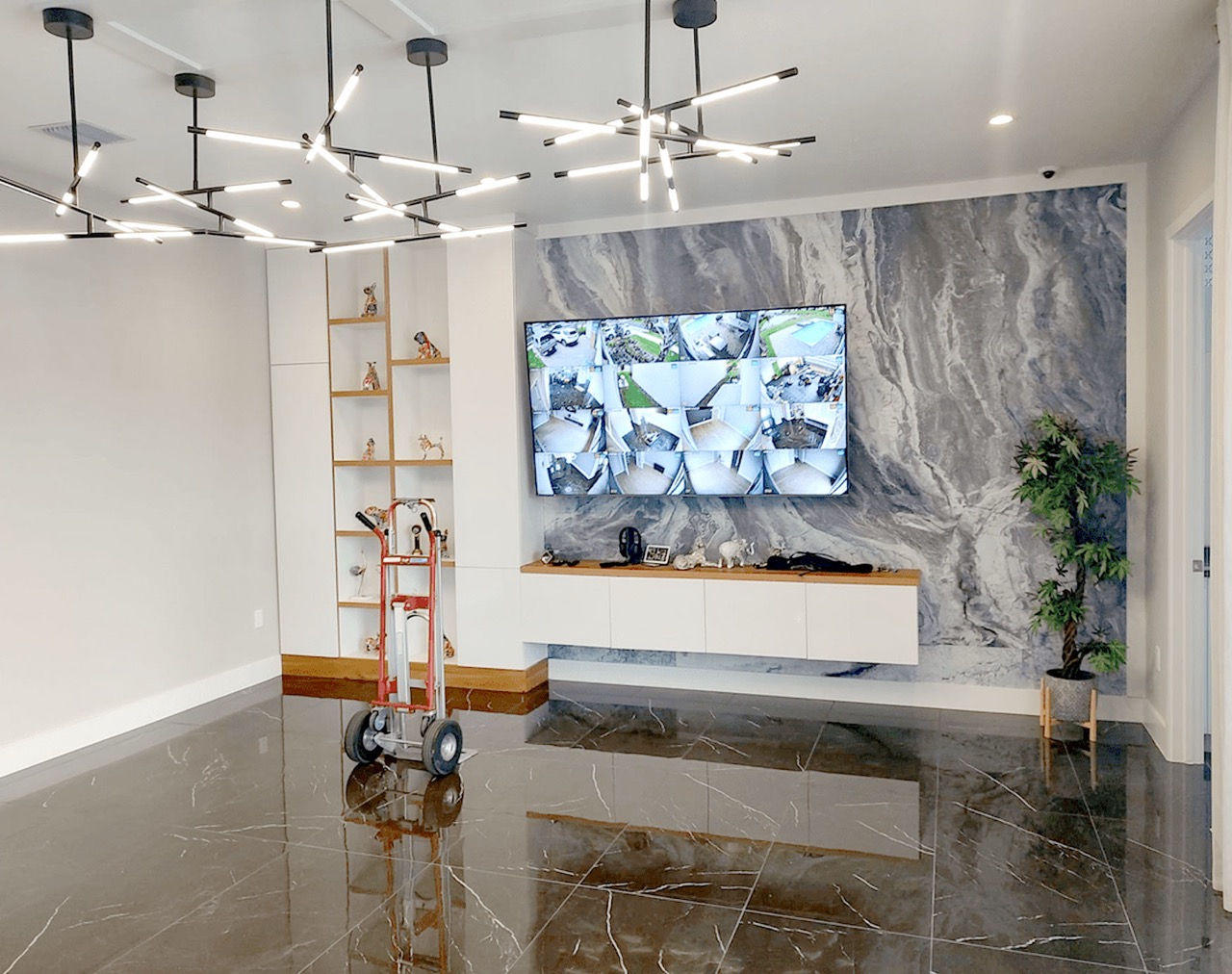 The image shows an interior space with modern decor, featuring marble flooring, a large stone wall with a television mounted on it, a contemporary light fixture hanging from the ceiling, and various pieces of furniture and decorative items.