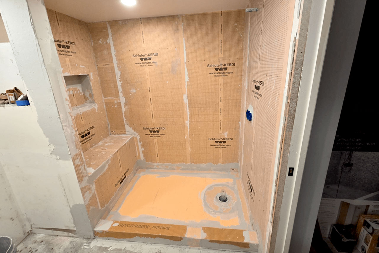 The image shows a bathroom under renovation with exposed walls and floor, featuring plywood and cardboard barriers, and a visible drainage system on the floor.