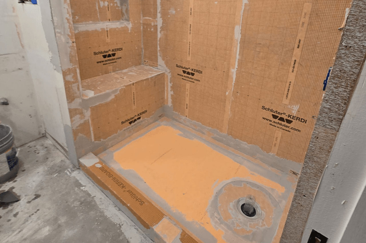 The image shows a bathroom under renovation with exposed walls and flooring, featuring a toilet without a tank, surrounded by construction materials such as plastic sheeting and cardboard.