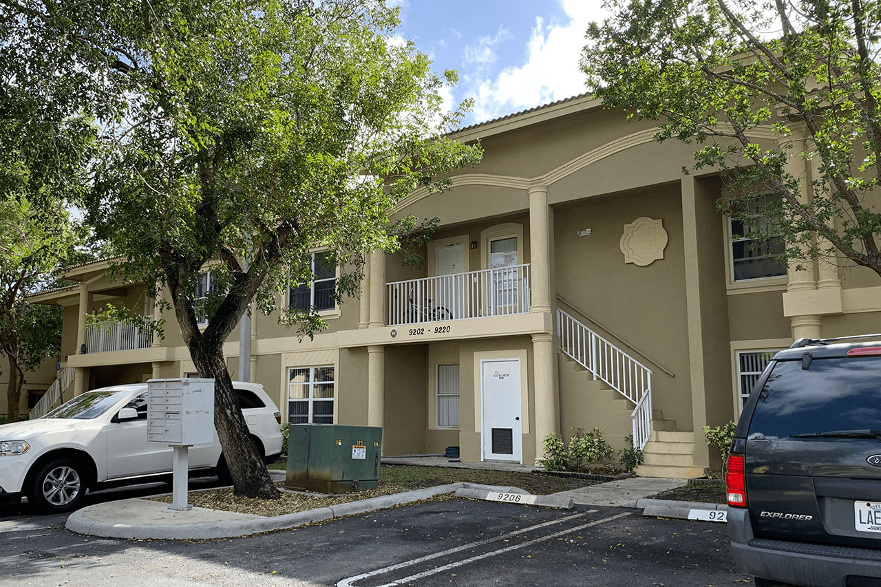 The image shows a multi-story residential building with a beige facade, a balcony on the second floor, and a white staircase leading up to it. The building has a sign that reads  123 Main Street  on its exterior wall. In front of the building, there is a parking lot with parked cars, and a sidewalk with trees lining it. The sky is clear, suggesting it might be a sunny day.