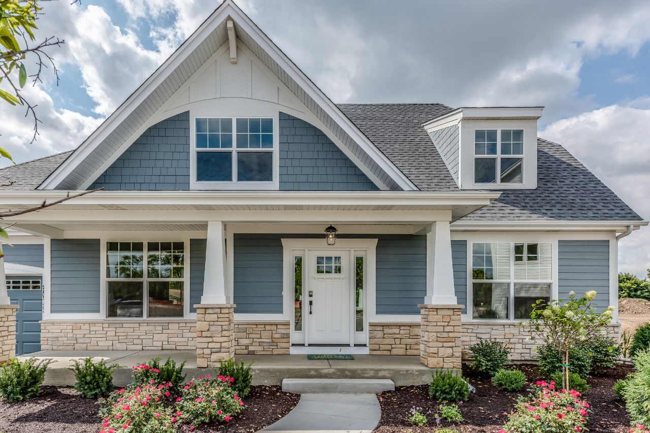 The image shows a two-story residential house with a blue exterior, white trim around the windows and doors, a gray roof, and a front yard landscaped with shrubs and a paved driveway.