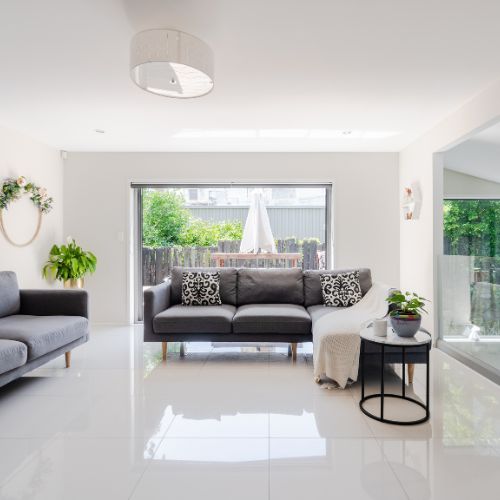 The image shows a modern living room with a white color scheme, featuring a large window revealing an outdoor patio area, a gray sectional sofa, a black coffee table, a white rug, a white wall-mounted light fixture, and a contemporary interior design style.