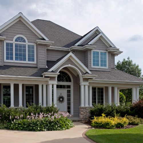 The image shows a two-story residential house with a white exterior, multiple windows, and a garage door, set against a landscaped yard with manicured grass and flower beds.