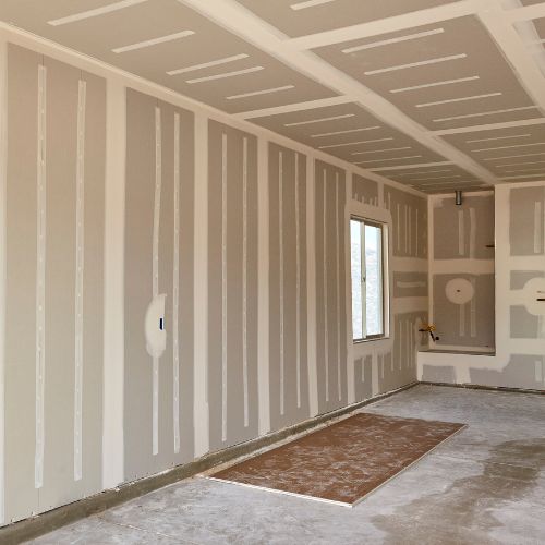 The image shows an interior view of a room under construction with unfinished walls and flooring, featuring insulation materials and exposed studs, alongside a partially framed doorway leading to another space.