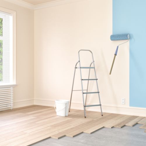 The image shows a room undergoing renovation with a ladder leaning against a wall, a paint roller hanging on a magnetic strip, and a blue wall corner with a white bucket placed on top of it.