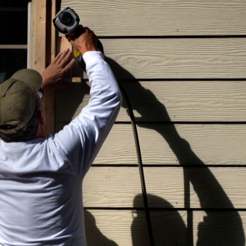 The image shows a person working on a house exterior, specifically installing siding, with a focus on a close-up action shot of spraying adhesive onto the wall.