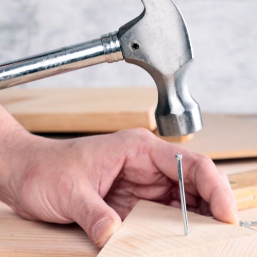 An individual is using a hammer to drive a nail into a wooden surface, with a focus on their hand and the tool they are using.