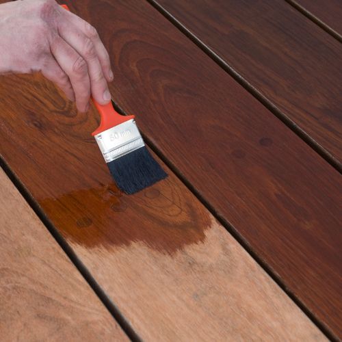 The image shows a person using a paintbrush with blue tape around its bristles to apply a dark brown wood stain on a wooden deck surface.