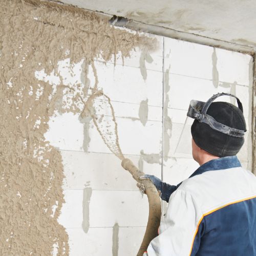 The image depicts a person engaged in wall repair work, using a trowel to apply a substance to a damaged section of a wall with visible peeling paint or plaster.