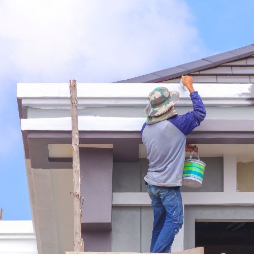 A construction worker applying paint to a house roof while standing on a ladder.