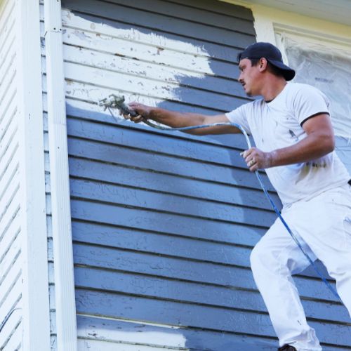 The image shows a person engaged in exterior house painting, using a sprayer to apply paint to a wooden surface.