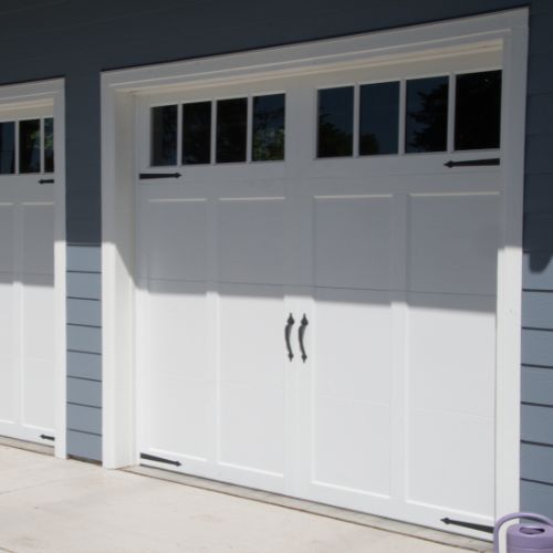 The image shows a section of a white garage door with a single window on the left side and a black handle on the right, set against a gray exterior wall of a house.