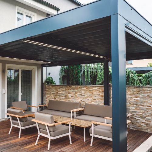 The image shows an outdoor patio area with a pergola structure featuring a black metal frame, a solid roof, and a brick wall behind it. Underneath the pergola, there are wooden benches and tables, with a brown sofa and matching chairs arranged for seating. The flooring is made of wood planks, and there s a glass sliding door leading to an indoor space. The patio is surrounded by a low stone wall, and the area is well-maintained with clean lines and minimal decoration.
