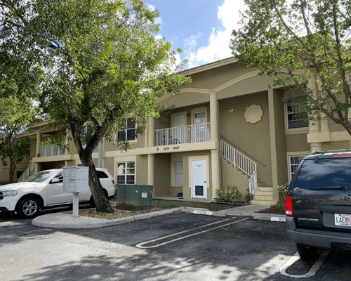 The image shows a two-story apartment building with a beige exterior during daylight hours under a clear sky, with palm trees visible in the background, and parked cars on the street in front of the building.
